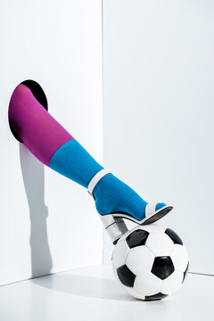 cropped image of girl putting leg in stylish violet tights, blue sock and white high heel on football ball through hole on white
