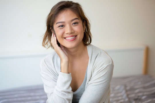 Portrait of cheerful young woman sitting on bed. Asian girl looking at camera and smiling indoors. Beauty concept