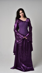 full length portrait of beautiful girl with long black hair,   wearing purple fantasy medieval gown. standing pose on grey studio background.