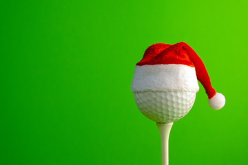 Golf ball in a red Santa Claus hat mounted on a tee. Sports concept on the theme of Christmas and New Year. Green background. Copy space.