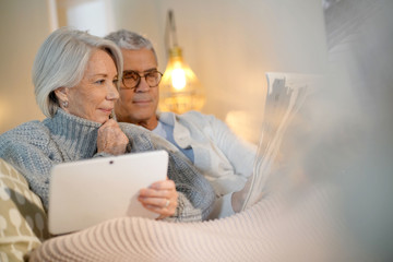  Senior couple relaxing at home on couch with tablet and newspaper