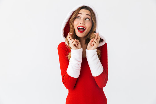 Image of joyful girl 20s wearing Christmas red dress keeping fingers crossed while standing, isolated over white background
