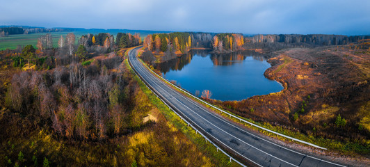 Aerial panorama of the asphalt road curving near the small lake with colorfull autumn trees on its coast