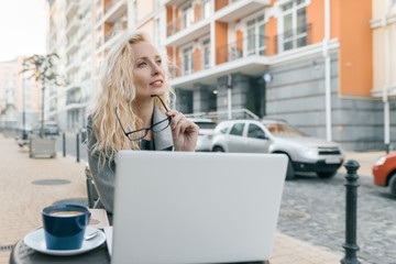 Portrait of young beautiful fashionable blond woman in warm clothes sitting in an outdoor cafe with laptop computer, drinking cup of coffee, city street background