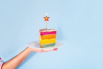 Female hand holding white plate with slice of Rainbow cake with birning candle in the shape of star isolated on blue background. Happy bithday, party concept. Square card. Selective focus. Copy space.