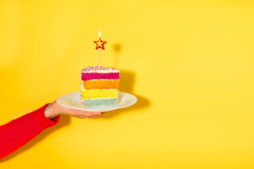 Female hand holding white plate with slice of Rainbow cake with birning candle in the shape of star...