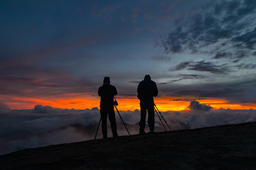 Two unidentified photographers take a photo of cloudy mountain landscape during sunrise.