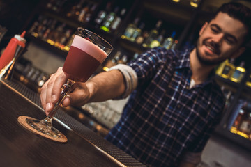 Young bartender standing at bar counter serving cocktail close-up cheerful