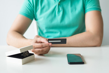 Closeup of buyer holding credit card. Person making online payment at table with smartphone and new watch in box. Online shopping and purchase concept. Cropped front view.