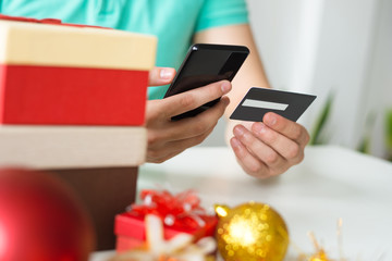 Closeup of man with credit card, smartphone and Christmas gifts. Person making online payment at table with small gift box. Christmas shopping concept. Cropped front view.