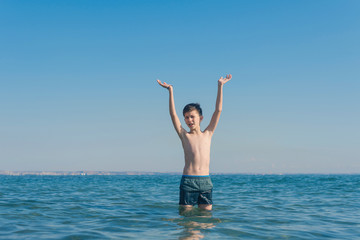 13 years old boy raises his hands standing up in  the sea waves. Concept of family summer vacation