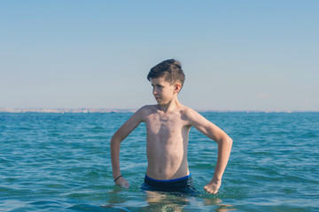 13 years old boy holding hands on hips and outraged standing in the sea waves. Concept