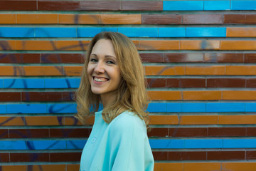 Close up portrait of smiling laughing young woman with fair wavy hair, wearing blue coat, near wall with colorful tiles, outdoors