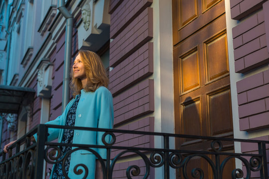 Beautiful smiling positive woman wearing dress and elegant blue coat is staying on doorstep, front porch of beautiful building with forged railings, sunbeam on face and door