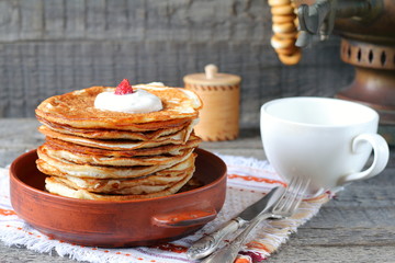 A stack of pancakes in a clay bowl near the old samovar on the gray wooden background.