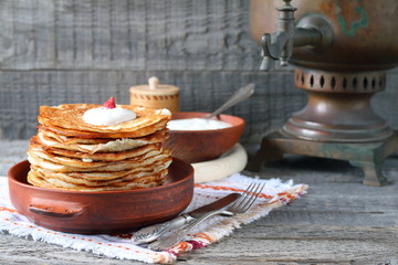 A stack of pancakes in a clay bowl near the old samovar on the gray wooden background.