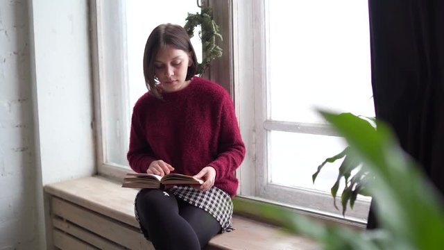 Pensive dreamy girl holding book sitting on sill at home looking at big window dreaming, thoughtful young woman thinking of reading new novel relaxing in modern cozy loft bedroom alone