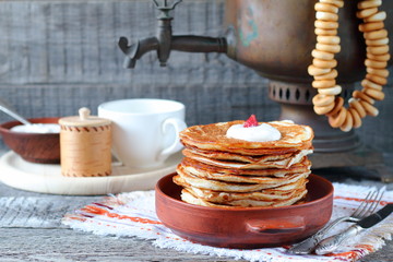 A stack of pancakes in a clay bowl near the old samovar on the gray wooden background.