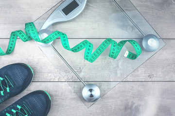 Green measure tape, glass weighing scale, running shoes on a rustic surface. Weight management and...