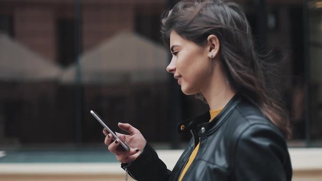 Young happy woman walking in the city street while using smartphone. Slow motion