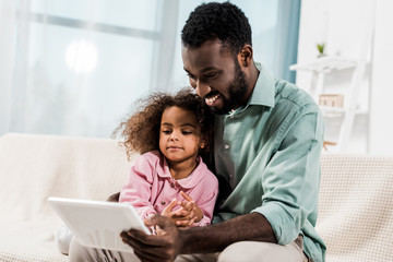 Obraz premium smiling african american father and kid using digital tablet while sitting on couch