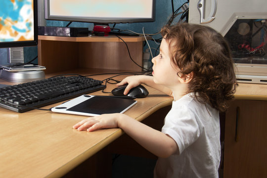 Little 2 3 year old baby girl in white clothers draws at the home computer in graphics drawing tablet. two monitors. The child is holding a mouse