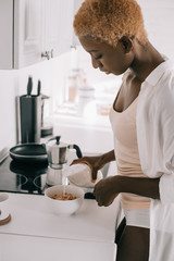 beautiful african american woman pouring milk in bowl with cornflakes in kitchen