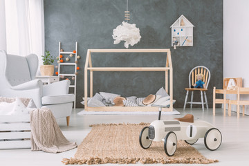 Lamp above wooden bed in grey baby's bedroom interior with car toy near armchair. Real photo