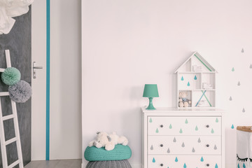 Green lamp on cabinet next to plush toy on pouf in white kid's room interior with ladder. Real photo
