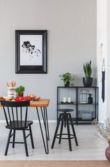 Black chair and stool at wooden table in grey dining room interior with plants and poster. Real...
