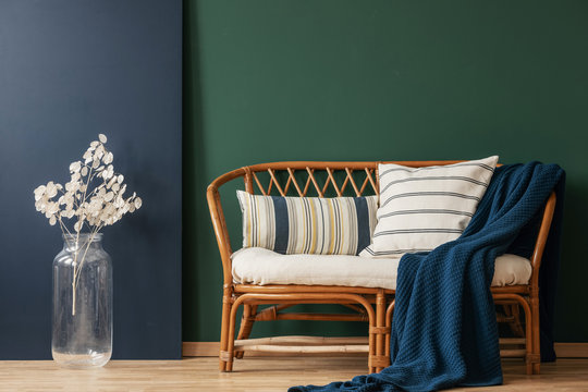 Natural wicker settee with striped pillows and blanket next to white flower in glass vase, real photo with copy space on empty green and blue wall