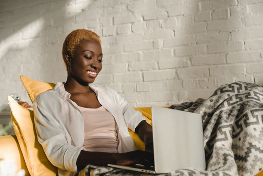 cheerful african american woman using laptop on yellow sofa