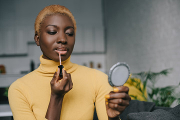 concentrated african american woman applying lip gloss and looking in mirror