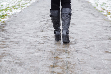 Young woman's legs in black leather boots walking on sidewalk in wet, warm winter day. Pavement...