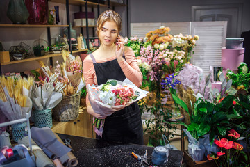 Busy young florist hold bouquet in hand. She talk on phone. Young woman stand in room full of flowers and plants.