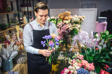 Cheerful young male florist hold colorful bouquet in hands. He look at it. Different plant and flowers are behind him. Guy stand inside.