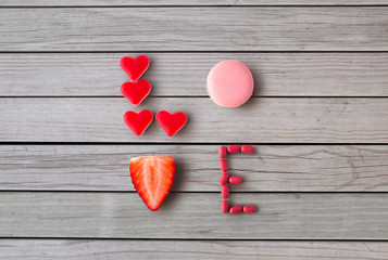 valentines day, sweets and confectionery concept - close up of word love made of red heart shaped candies, pink macaron cookie and strawberry over grey wooden boards background