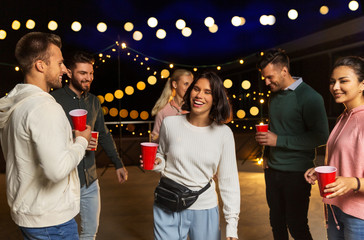leisure, celebration and people concept - happy friends with drinks dancing at rooftop party at...