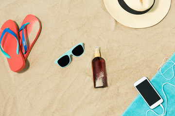 vacation and summer holidays concept - smartphone with earphones on towel, straw hat, sunglasses, flip flops and bottle of sunscreen oil on beach sand