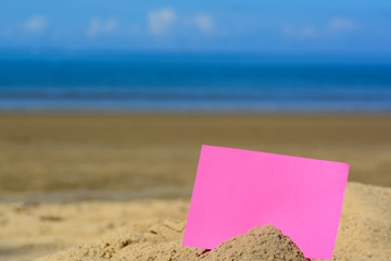 A blank red card buried in sand at the beach with blue ocean and blue sky.