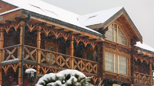Close view of cottage with mezzanine in mountain village during heavy snowfall with russian style carved wooden facade. Snow falling on log chalets at ski resort. Cold frosty winter day. Slow motion.