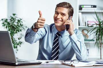 Cheerful businessman talking on the phone and showing thumb up. Photo of successful man working in...