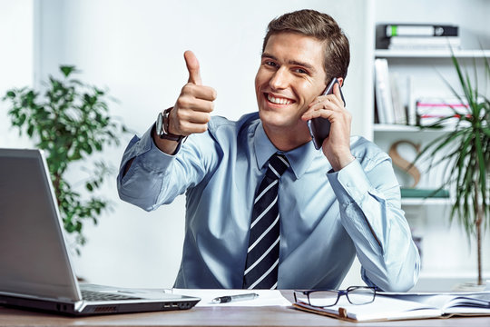 Cheerful businessman talking on the phone and showing thumb up. Photo of successful man working in the office. Business concept