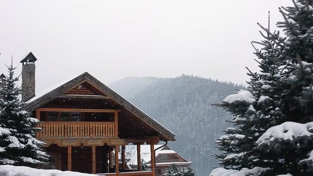 Christmas wooden mansion in mountains on snowfall winter day. Cozy chalet on ski resort near pine forest. Cottage of round timber with wooden balcony. Fir-trees covered with snow. Chimneys of stone.