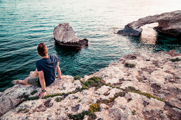 Young man sitting at the Love Rock in Cyprus, Greece watching the sunset.