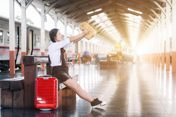 Asian pregnant woman traveler sitting on howing a Teddy Bear with a red suitcase in railway station travel.