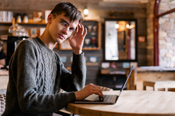 Stylish guy with glasses sitting in a cozy cafe at the laptop typing looks towards the camera