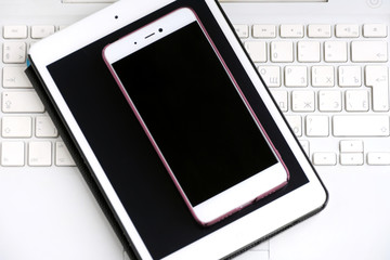 Tablet and mobile phone with empty black screens on the white laptop keyboard. Electronic touch device with selective focus on pink background. Mobility web office. 