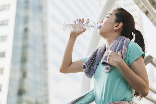 Happy beautiful young Asian woman drinking water after workout exercising on morning summer in city outdoor portrait. Asian female model holding bottle water in her hand. lifestyle, ,health concept