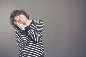 Young man in striped black and white sweater wants to sleep. Put his head in his hands and closed eyes while smiling. Standing on the grey background. Hands under his cheek. Insomnia concept